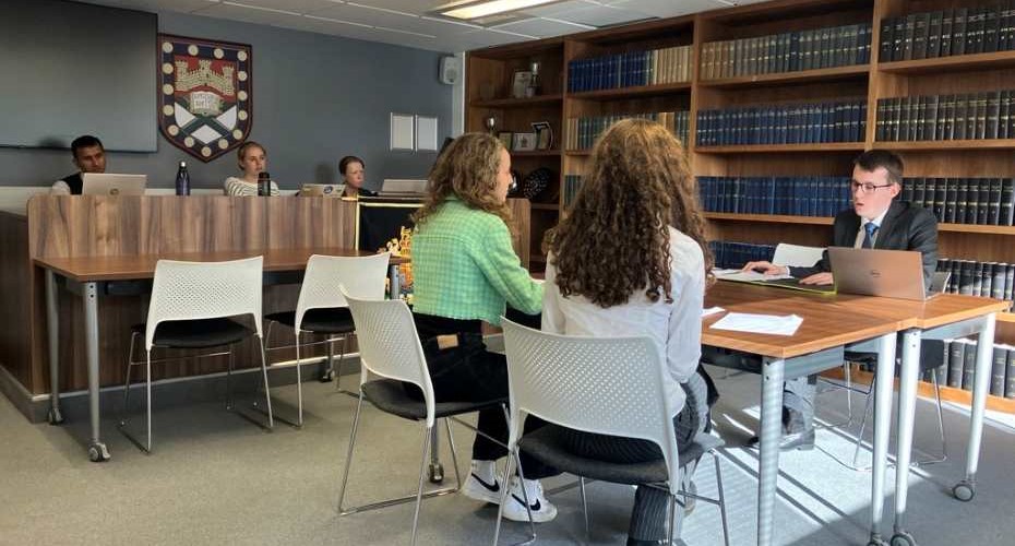 A mock court room, with tables and chairs, bookcases and the University shield on the wall. At one table there are two students facing a client. To the left are three observers to the conversation taking place.
