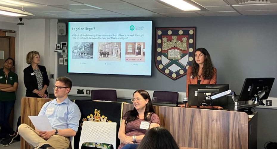 Four people in the Moot Court, slide presentation in the background. University of Exeter shield on the wall